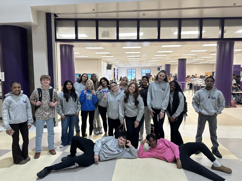 The same group of high school students stand together in a cafeteria entrance area, smiling and posing for a photo. Many are dressed in matching gray hoodies with a purple logo, and the busy cafeteria with students and serving lines can be seen behind them.