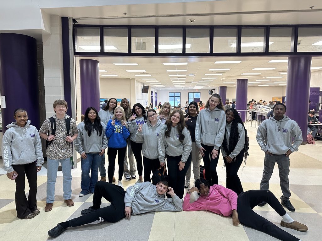 A group of high school students pose together in a school cafeteria area with purple columns and bright overhead lighting. Most are wearing matching gray hoodies with a purple logo, while two students lie playfully on the floor in front of the group. Other students and lunch tables are visible in the background.