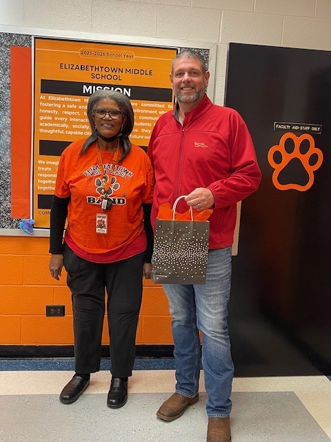 A bus driver wearing an orange Elizabethtown Middle School shirt stands indoors beside a man in a red jacket. They are smiling and holding a gift bag in front of a wall display featuring the school’s mission statement and a large Cougar paw print.