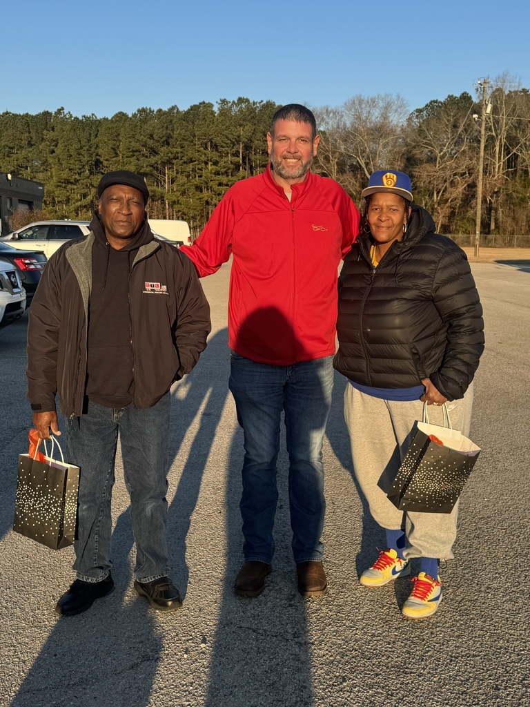 Two bus drivers stand on either side of a man in a red jacket in the school parking lot. Both drivers are holding appreciation gift bags. Trees and parked vehicles are visible in the background in warm morning sunlight.
