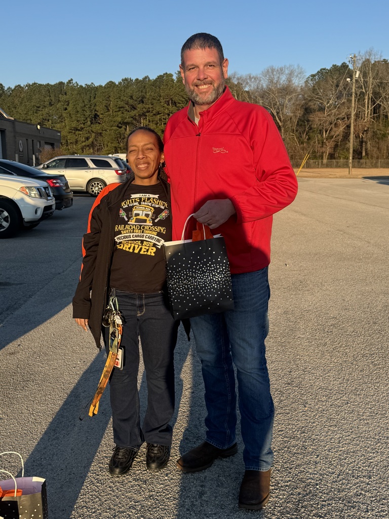 A smiling female bus driver wearing a “School Bus Driver” shirt stands in a school parking lot beside a man in a red jacket. They are holding a small appreciation gift bag. Parked cars and trees are visible in the background during early morning light.