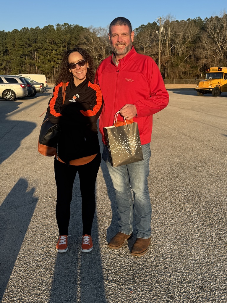 A bus driver wearing black and orange Cougar gear and sunglasses stands next to a man in a red jacket in the school parking lot. A yellow school bus is visible in the background. They are smiling and holding a small appreciation gift bag.