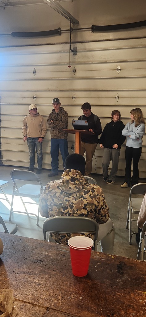 Five FFA members stand behind a wooden podium in a garage-style classroom, with one student presenting from a laptop while others observe and smile.