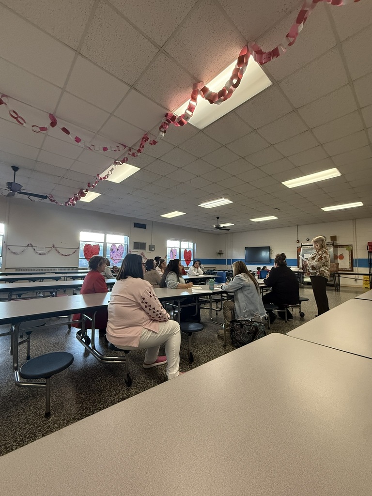 Parents and staff members sit together at cafeteria tables during a school meeting while a woman stands at the front speaking to the group. Pink and red paper chain decorations hang from the ceiling, and heart artwork is displayed on the windows. The setting appears to be a Parent Advisory Meeting or school event held in the cafeteria.