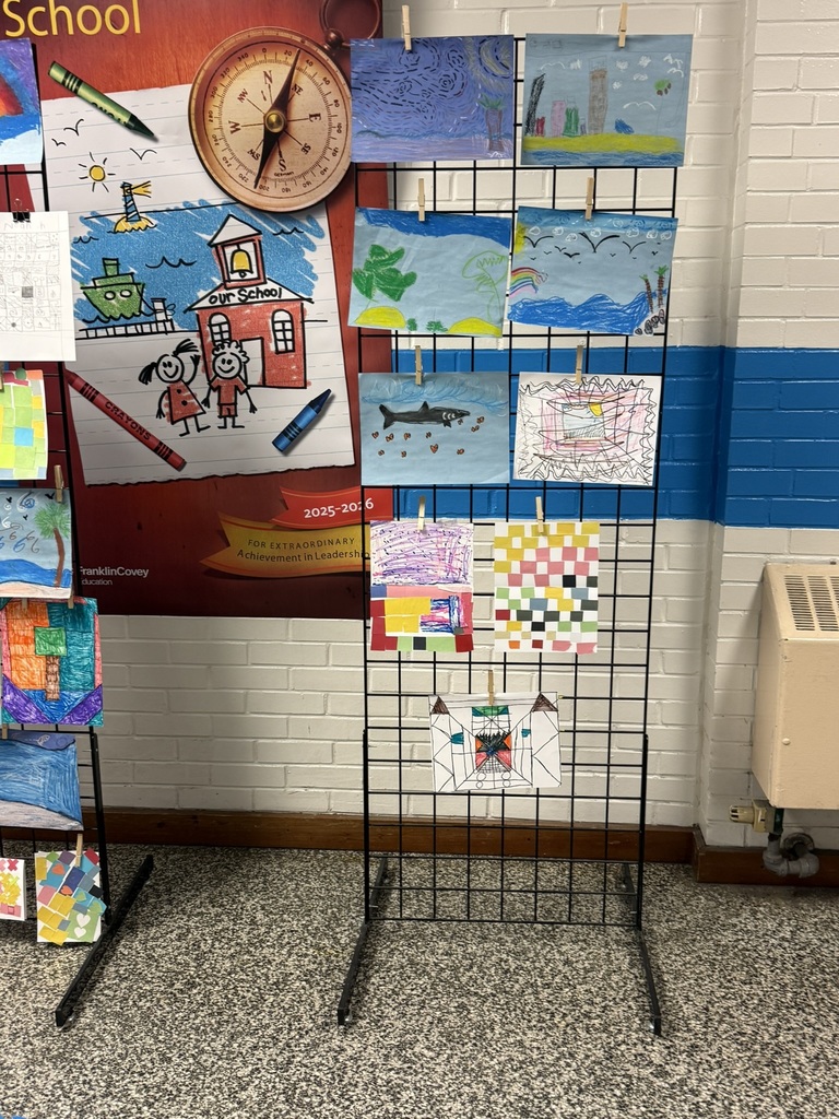 A black metal grid display stand holds several student drawings clipped in rows. The artwork includes colorful crayon and watercolor scenes such as ocean waves, sharks, palm trees, city skylines, abstract patterns, and geometric designs. The display is set against a white brick and blue painted hallway wall near a “Leader in Me School” poster.