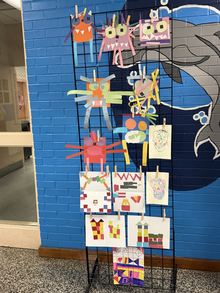 A tall grid display stand features vibrant paper monster art created from colorful construction paper. Each monster has unique shapes, bold colors, googly-style eyes, and creative details like long arms, teeth, and textured strips. The display stands against a blue and white brick wall with a dolphin mural partially visible behind it.