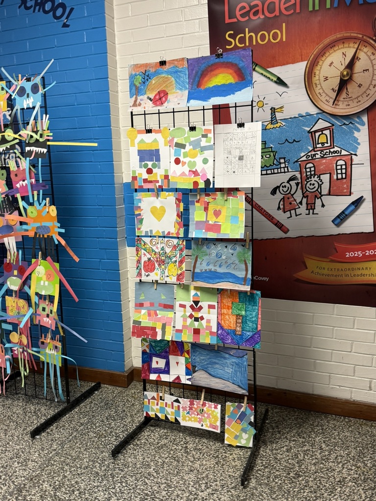 Another black grid display stand showcases a variety of student artwork, including bright mosaic-style paper collages, heart-themed designs, sunsets, rainbows, landscapes, and patterned geometric art. The artwork is clipped neatly in rows and displayed in the school hallway beside a large school-themed poster.