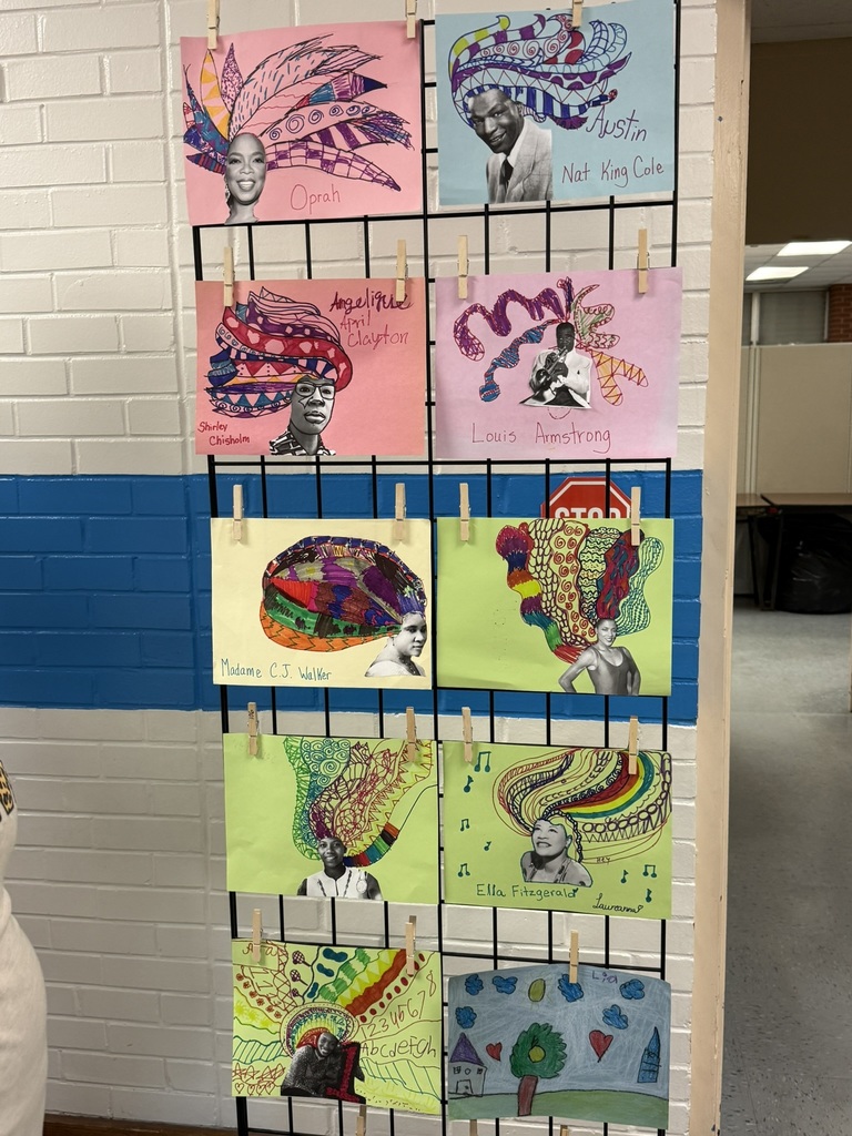 A tall grid display stand features vibrant paper monster art created from colorful construction paper. Each monster has unique shapes, bold colors, googly-style eyes, and creative details like long arms, teeth, and textured strips. The display stands against a blue and white brick wall with a dolphin mural partially visible behind it.