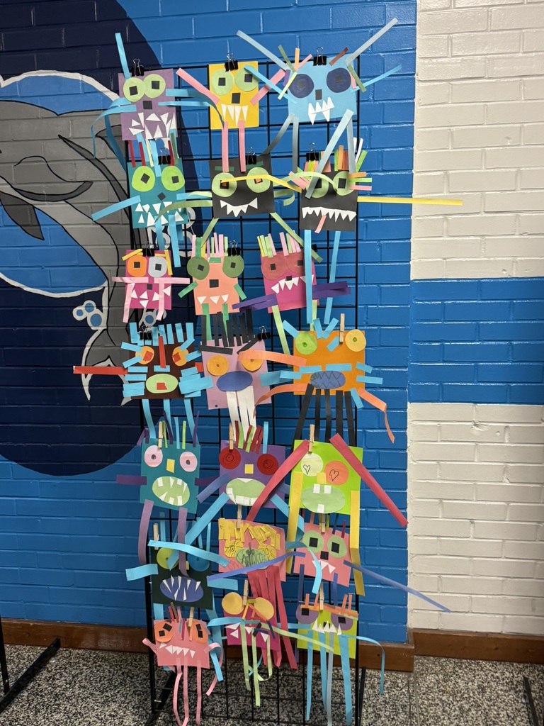 A tall grid display stand features vibrant paper monster art created from colorful construction paper. Each monster has unique shapes, bold colors, googly-style eyes, and creative details like long arms, teeth, and textured strips. The display stands against a blue and white brick wall with a dolphin mural partially visible behind it.
