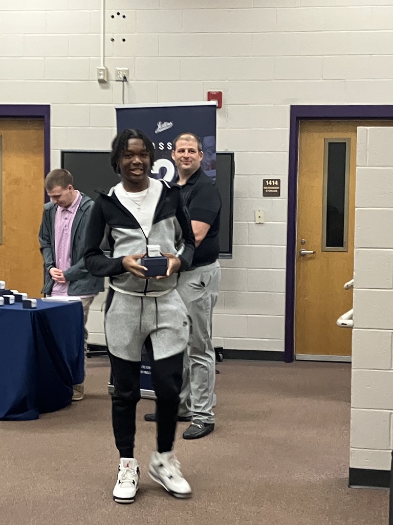 A young man in a grey and black tracksuit stands smiling while holding a small blue jewelry box. He is being presented with the award by a man in a black polo shirt, with another man in a pink shirt standing behind a table in the background.