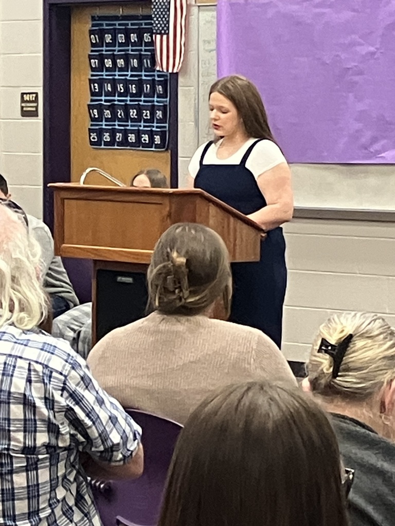 A young woman stands at a wooden podium speaking to an audience. Behind her is a purple wall with a large 'Attendance Matters' calendar and an American flag.