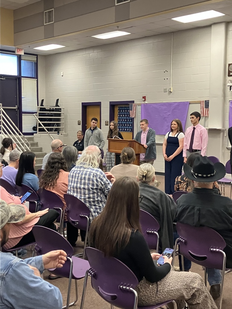 A group of five high school students stands behind a wooden podium during a presentation in a school media center. A crowd of seated adults and students face them, with an American flag and purple wall decorations in the background.