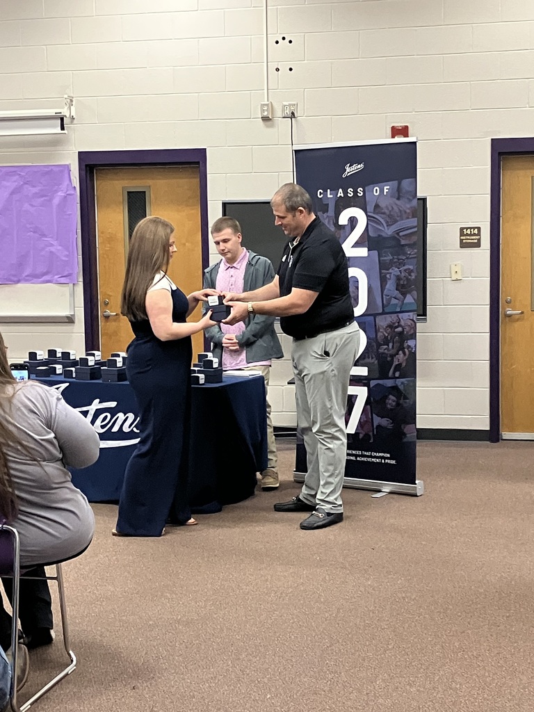 A high school student in a graduation gown receives a small gift box from a man in a black polo shirt during an indoor ceremony. Another student stands nearby against a purple and white wall with a 'Class of 2024' banner in the background.
