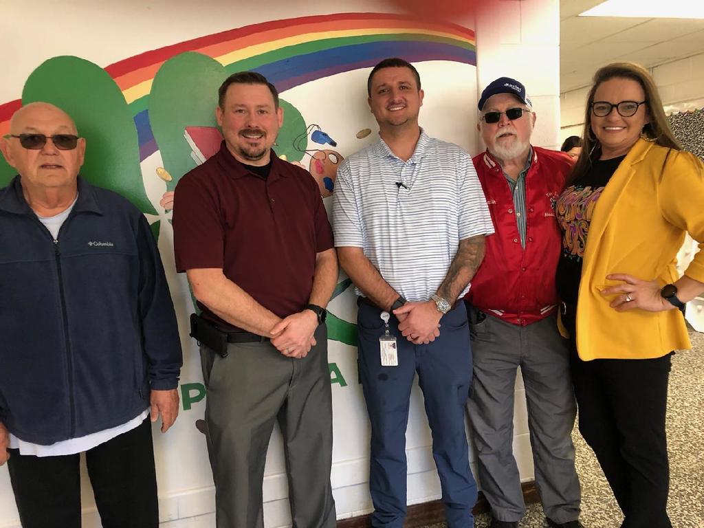 Alt text: Five adults stand side by side indoors in front of a colorful wall mural featuring a rainbow and green shapes. Three men and one older man wear casual or business-casual clothing, including polos and jackets; one older man wears sunglasses and a blue cap. A woman on the far right wears glasses, a black outfit with a bright yellow blazer, and smiles with her hand on her hip. The group appears to be posing together in a school hallway.