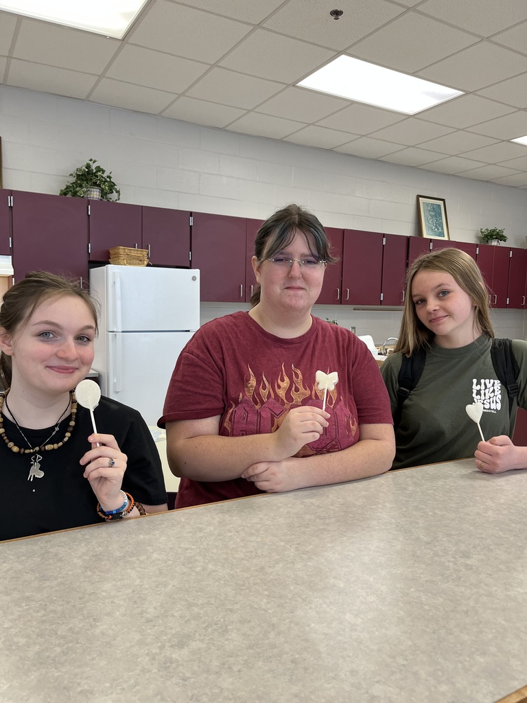 Three students in a classroom kitchen smiling and holding up their handmade lollipops.