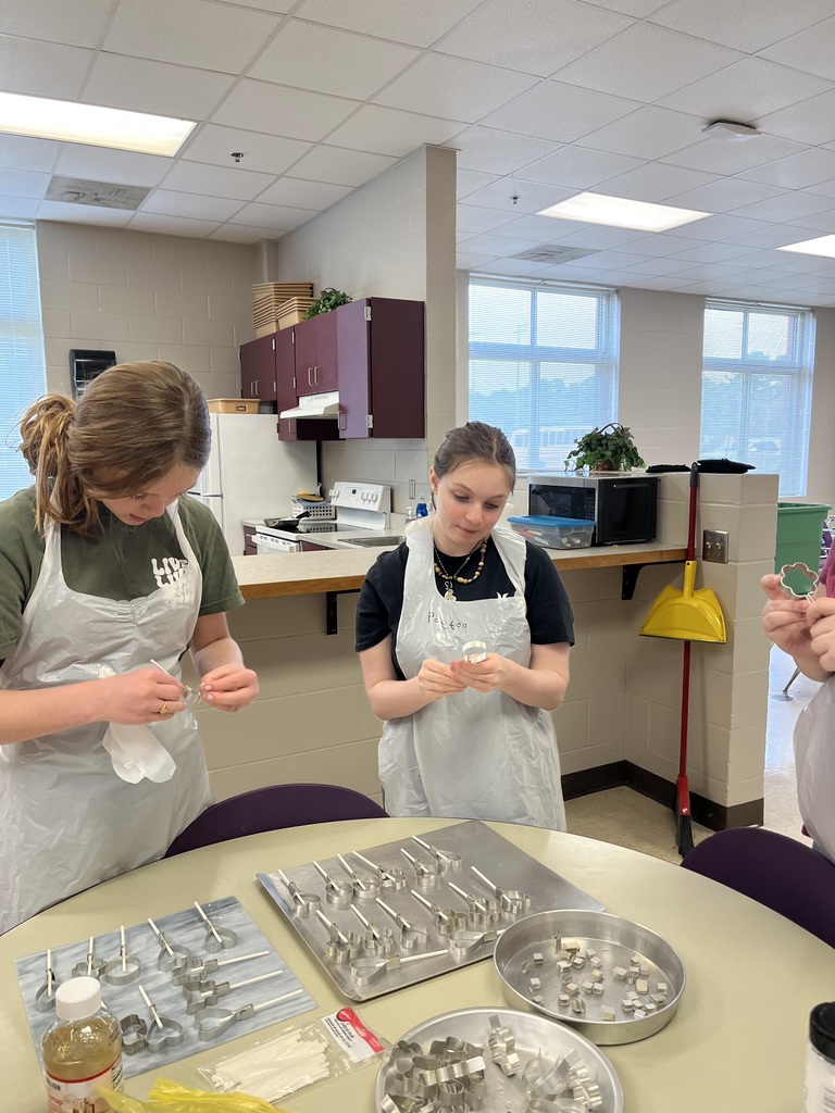 Two students in aprons and hairnets at a table, carefully making lollipops using metal molds in a classroom kitchen.