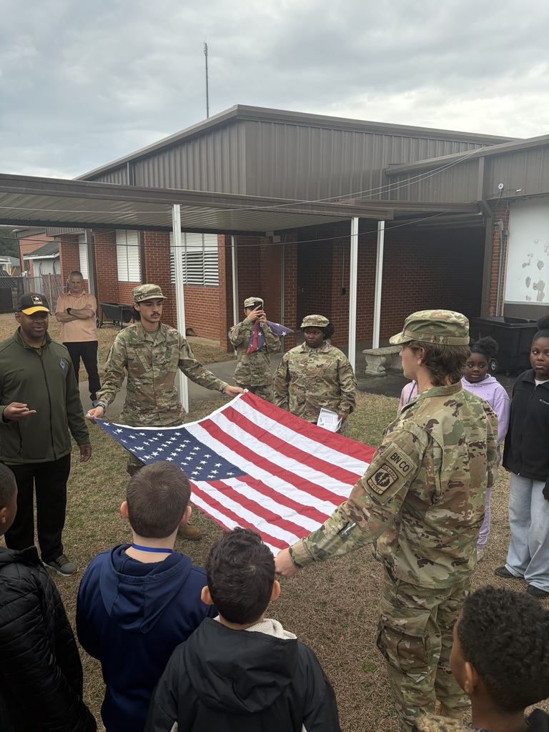 A JROTC student and a school staff member hold the American flag while explaining the folding process to students gathered around them.