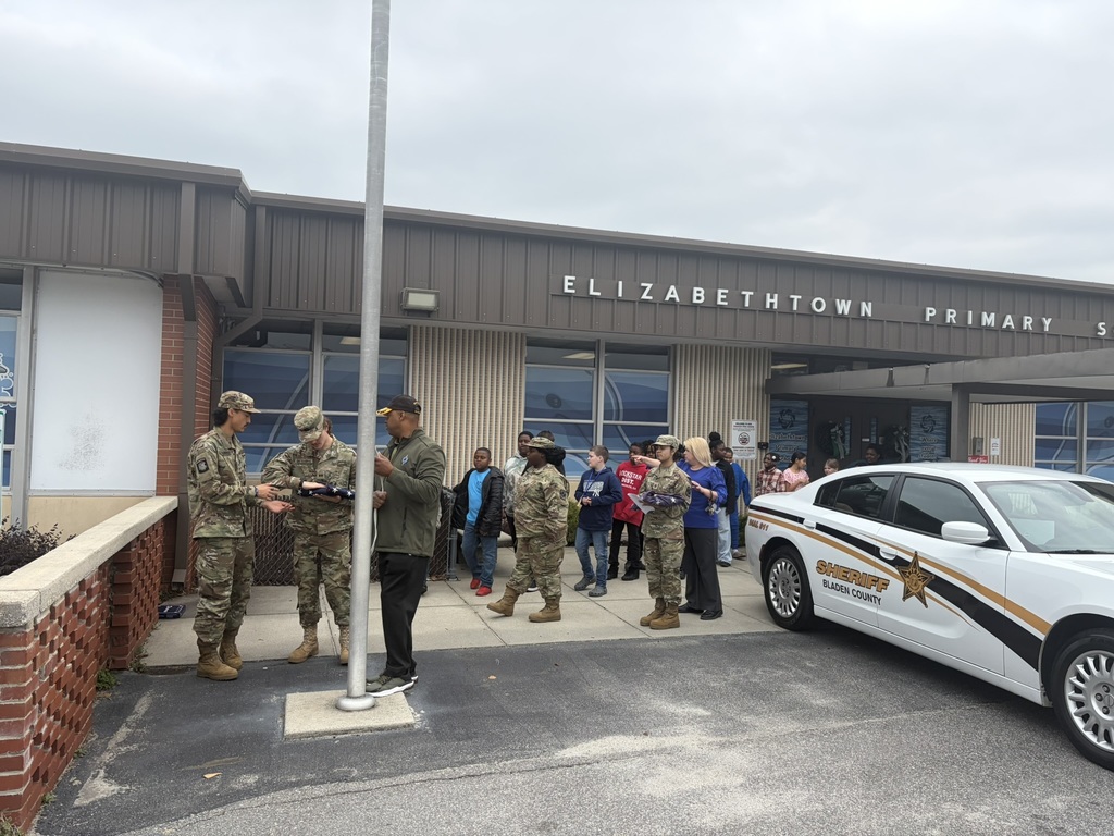 JROTC students and a school resource officer stand near the flagpole in front of Elizabethtown Primary School preparing for the flag folding demonstration.