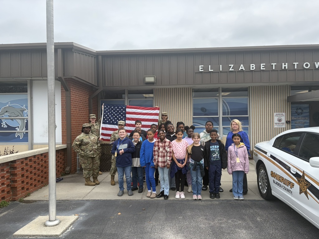 A group photo in front of Elizabethtown Primary School shows fourth graders and JROTC students smiling together, with the American flag displayed and a Bladen County Sheriff’s vehicle parked nearby.