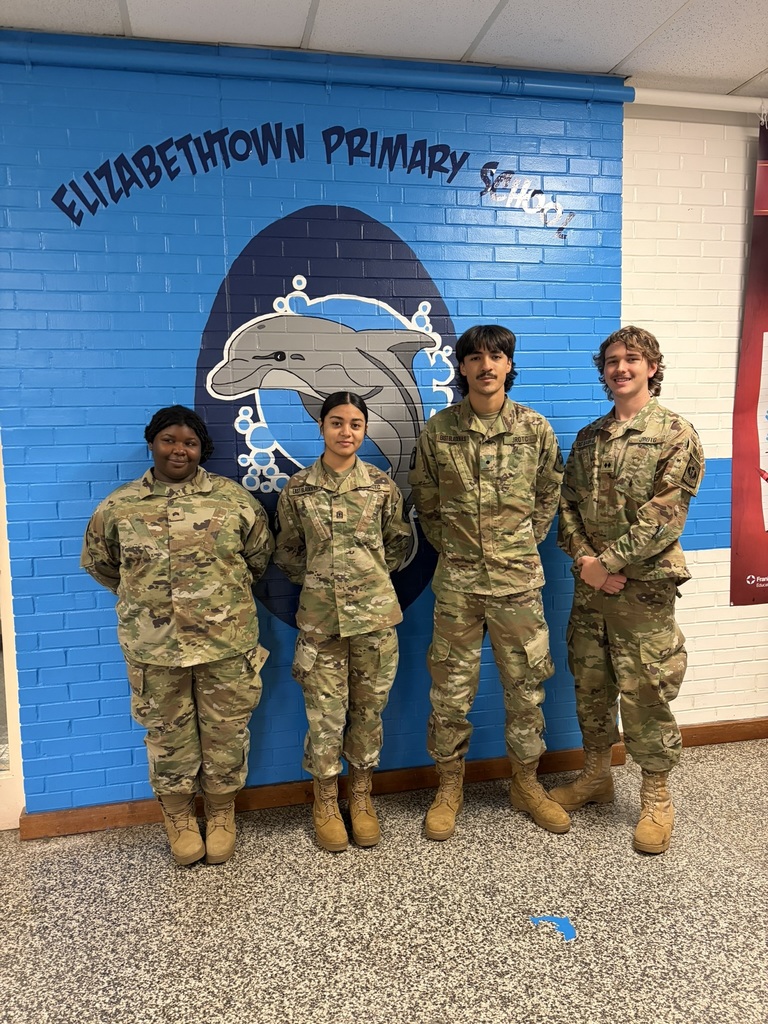 Four high school JROTC students in camouflage uniforms stand in front of the Elizabethtown Primary School dolphin mural inside the building.