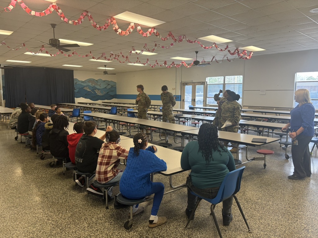 JROTC students speak to a group of fourth graders seated at cafeteria tables while a staff member stands nearby.