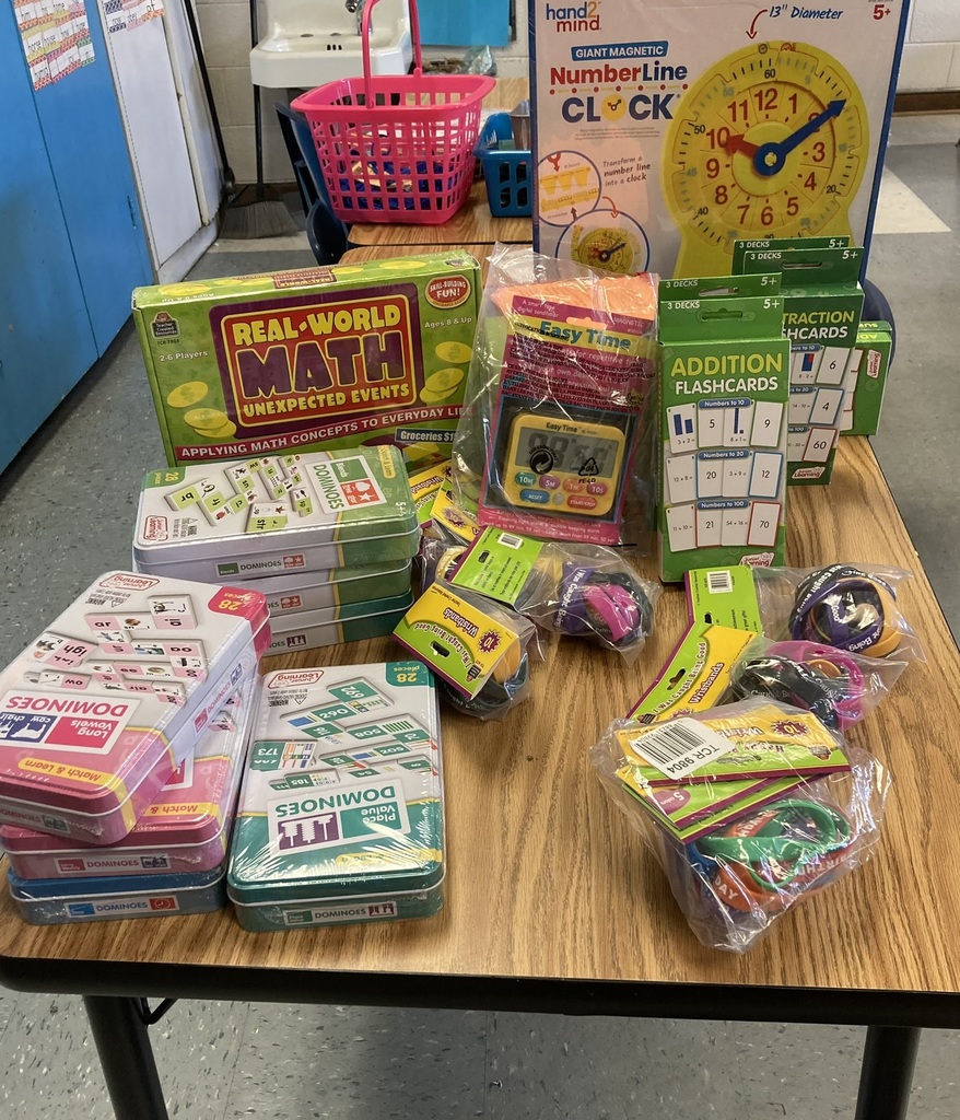 A classroom table filled with new educational materials for second grade, including math domino sets in metal tins, a Real-World Math game, addition and fraction flashcards, a giant magnetic number line clock, timers, and small math manipulatives in plastic packaging. A pink basket and classroom furniture are visible in the background.