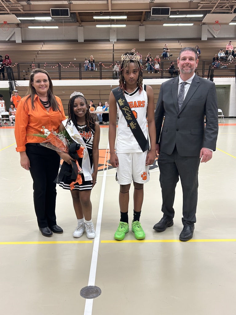 Four individuals stand together at center court in a school gymnasium during a basketball event. Two students wear crowns and sashes, suggesting a homecoming or court recognition. The female student is dressed in a black and white cheer uniform and holds a bouquet of flowers. The male student wears a white “Cougars” basketball uniform with a black sash and crown. They are flanked by two adults—one woman in an orange blouse and black pants, and one man in a gray suit and tie. Bleachers with spectators are visible in the background, along with orange and black school decorations.