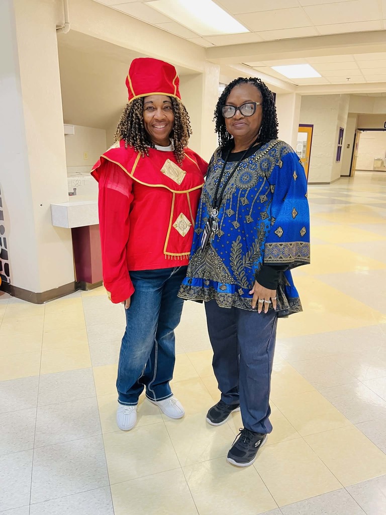 Two smiling Black women standing in a school hallway. The woman on the left is wearing a bright red academic or ceremonial robe with gold trim and a matching headpiece. The woman on the right is wearing a blue and black patterned dashiki-style top and glasses.