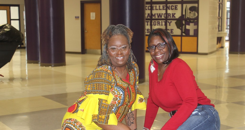 Two women smiling in a school hallway; one wears a yellow and black patterned dashiki with glasses, and the other wears a bright red long-sleeved top.