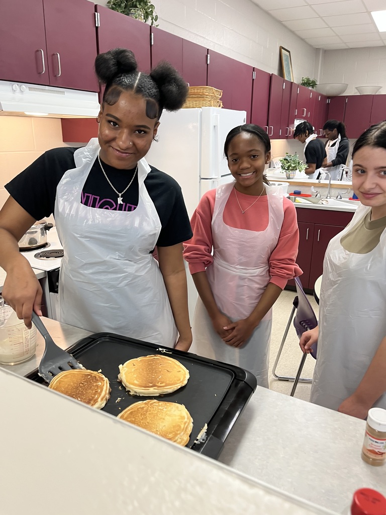 Three smiling students pose next to a griddle with pancakes
