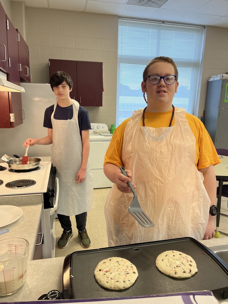 One boy stands next to a stove stirring a pan while a second boy stands next to a griddle with a spatula before flipping pancakes