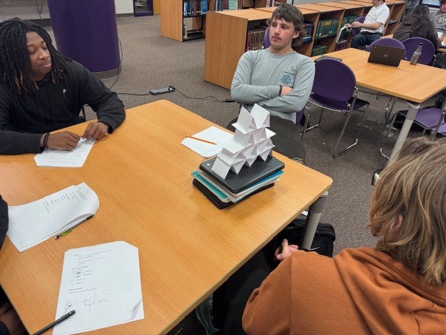 Two students sit at a wooden table in a classroom; one works on a paper while the other watches. A tall, white geometric paper sculpture sits on a stack of books between them