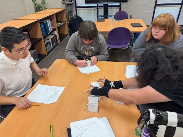 Four students sit around a library table collaborating on a project. They are focused on arranging small white rectangular blocks and writing on papers. More students are visible in the background.