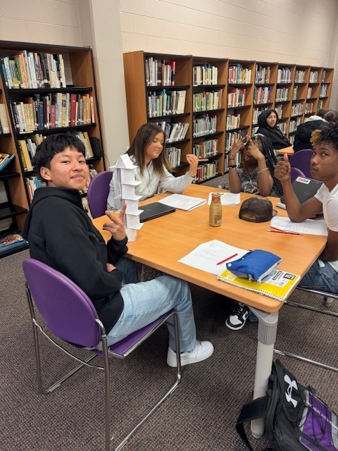 Four high school students smiling and collaborating on a paper tower project at a table in a library with bookshelves in the background.