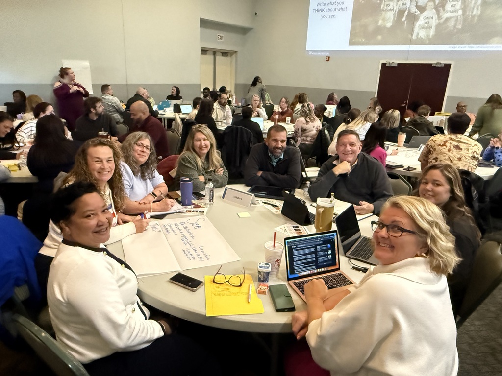 Large group of educators seated at round tables in a conference room, smiling and collaborating during a professional development session with laptops, notes, and chart paper visible.