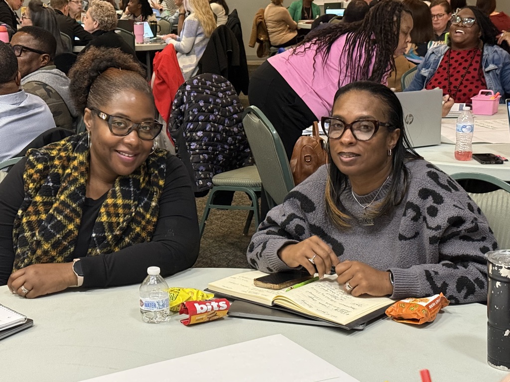 Two educators seated at a round table with notebooks and snacks, smiling toward the camera during a collaborative training session in a busy conference space.