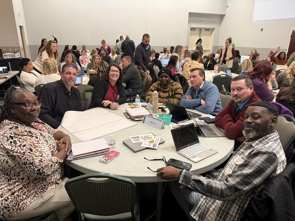 Team of educators gathered around a round table with laptops and materials, smiling and working together during a professional learning workshop in a conference room.