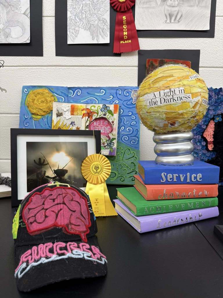 Art display featuring a yellow paper globe labeled “A Light in the Darkness,” stacked books labeled Service, Character, Achievement, Leadership, and a yellow third-place ribbon.