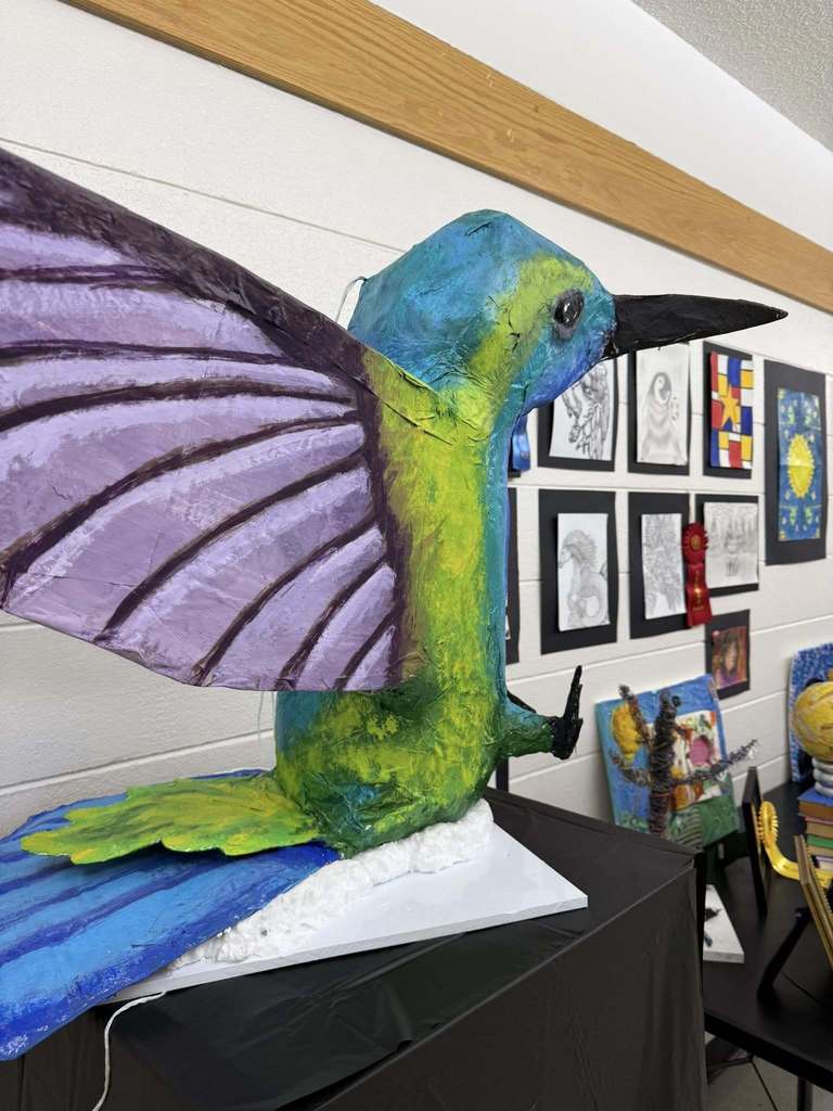 Large colorful hummingbird sculpture with purple wings and green-blue body perched on a base, with student artwork displayed in the background.