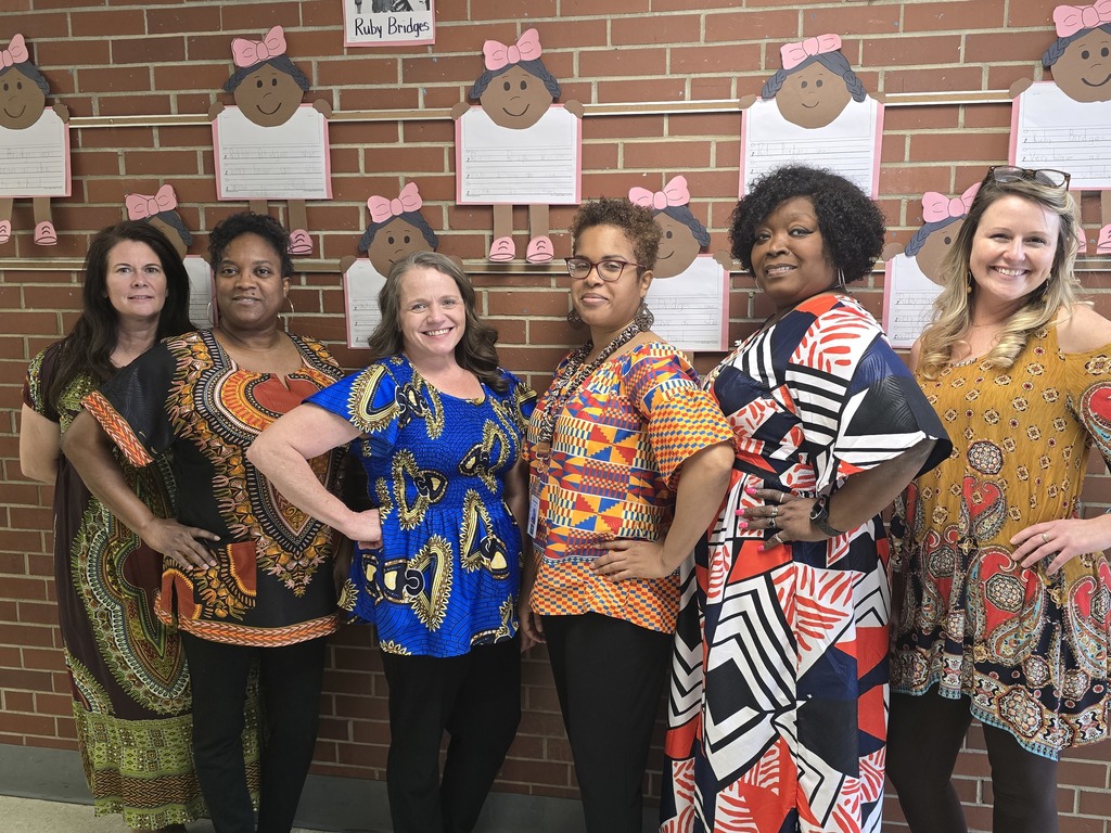 Six staff members stand together smiling in a school hallway, dressed in colorful cultural attire to celebrate Black History Month. Behind them, student artwork inspired by Ruby Bridges is displayed on a brick wall, highlighting student learning about influential Black leaders.