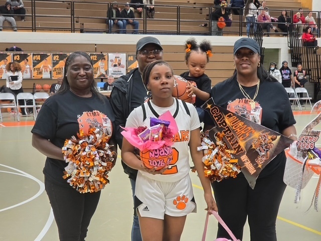 ETMS girls basketball player stands with family members holding orange and black pom-poms, balloons, and a senior night gift basket during recognition on the gym floor