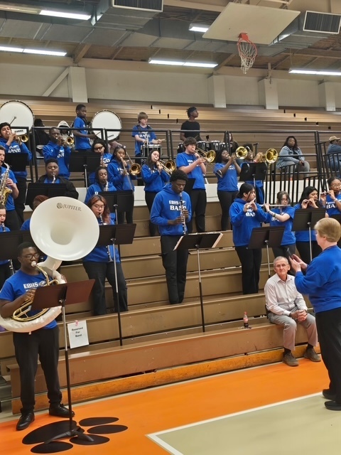 High school band performing in the gym bleachers. Students in blue band shirts play brass and woodwind instruments while a director conducts in front. Spectators sit in the background.
