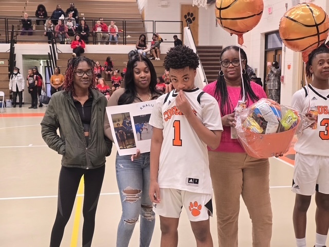 ETMS basketball player wearing jersey number 1 stands with family members holding a framed photo collage and gift basket. Basketball balloons float above them in the gym.