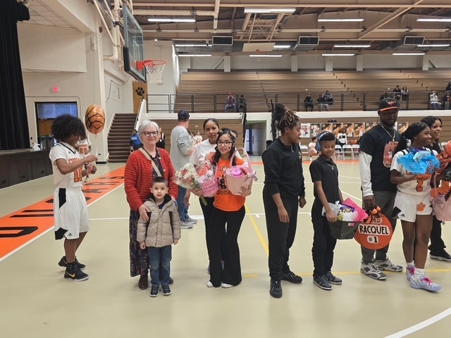 Basketball player stands on the court with family members during a senior night recognition. Family members hold flowers and gift baskets while spectators watch from the bleachers.