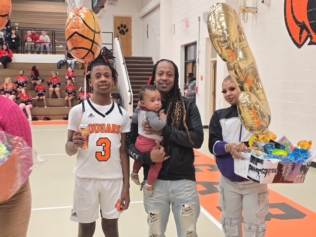 Cougars basketball player wearing jersey number 3 stands with family members during senior night recognition. One family member holds a baby while another holds a large gold balloon and gift basket.