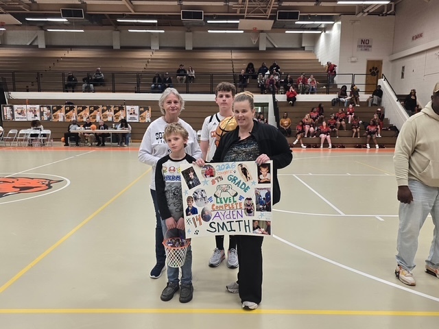 Student athlete stands with family members holding a decorated senior poster board that reads “8th Grade Level Complete.” They pose together on the gym floor during recognition.
