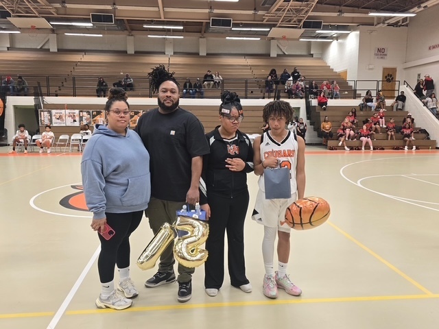 ETMS basketball player stands with family members holding gold number balloons and gifts during senior night recognition. Cheerleaders and spectators sit in the bleachers behind them.