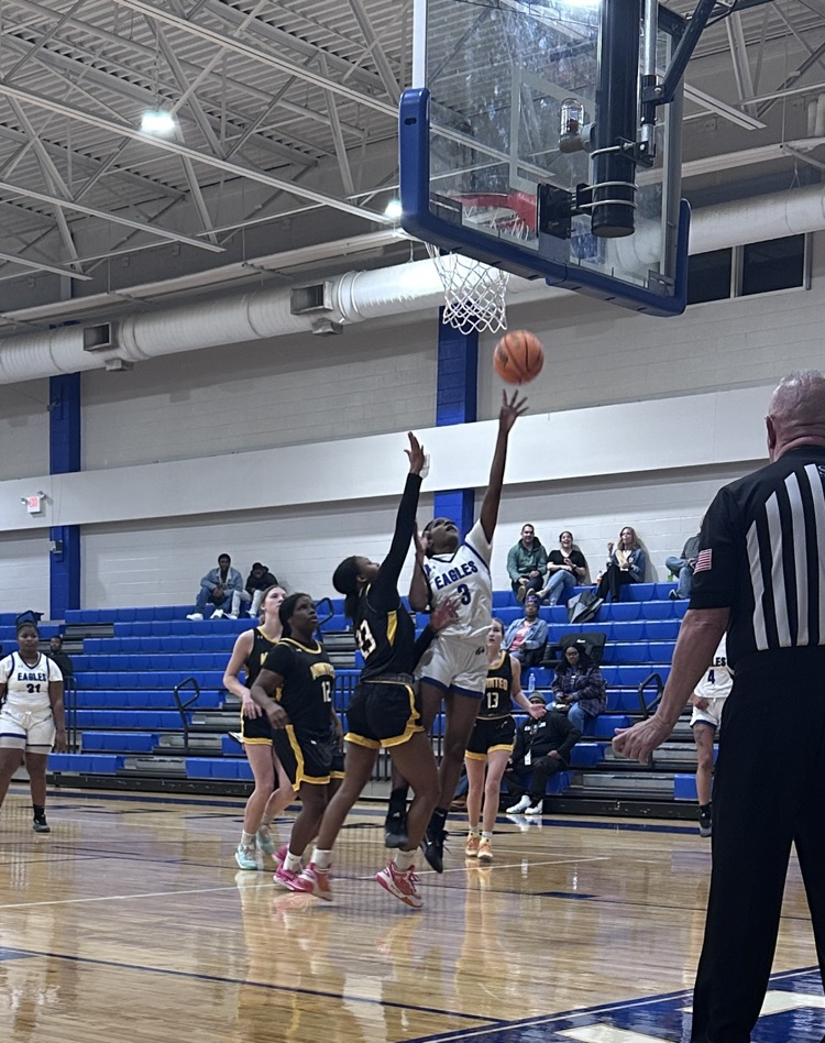 A girls basketball player in a white Eagles jersey drives to the basket for a layup as two defenders in black and gold attempt to block the shot. The referee stands nearby while spectators watch from blue bleachers in the background.