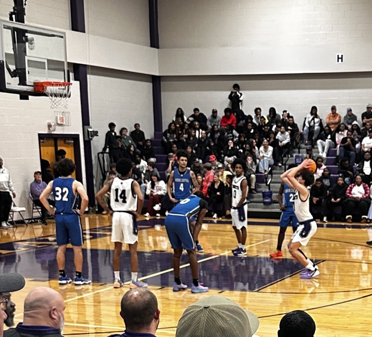 A boys basketball player in a white jersey prepares to shoot a free throw while players from both teams line the lane. A large crowd fills the bleachers behind them in a packed high school gym.