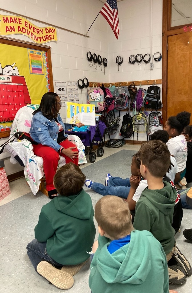 A teacher reads a Dr. Seuss book aloud to a group of elementary students seated on the floor in front of her. The students listen attentively in a classroom decorated with bulletin boards, backpacks hanging on hooks, and an American flag on the wall.